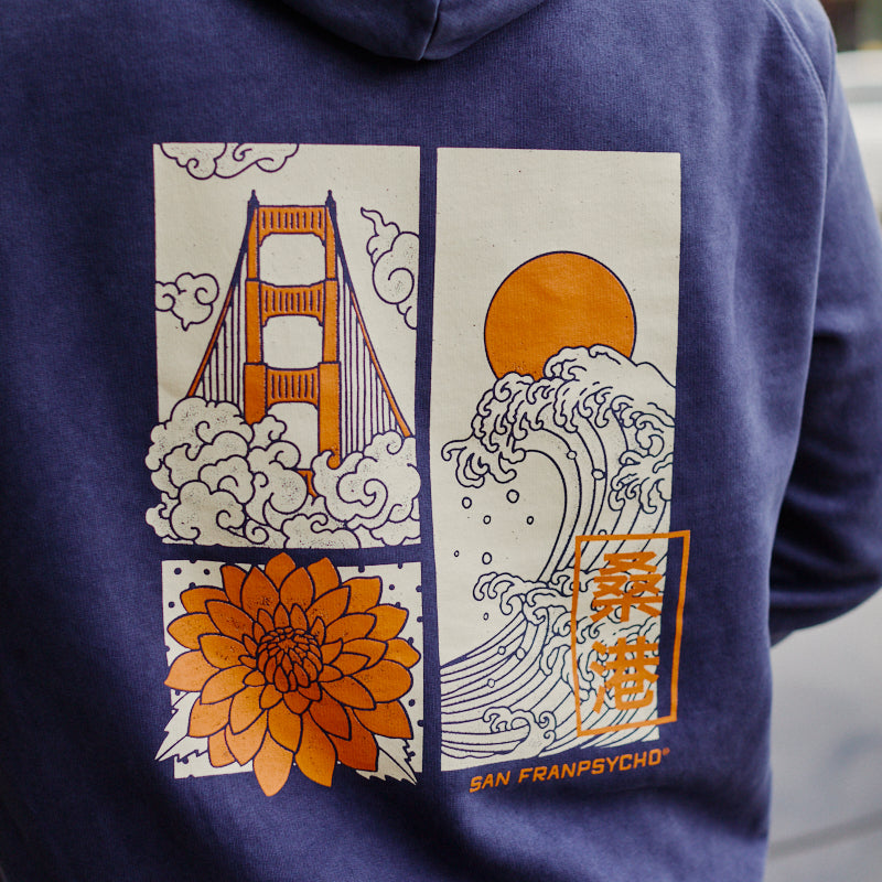Wearing the Sunset Dahlia Pullover Hoodie featuring hand-printed artwork of a bridge, flower, waves, and the sun on the back—crafted in San Francisco for an authentic touch.