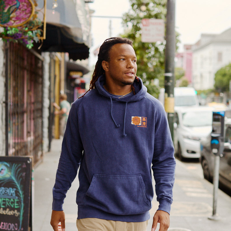 A man in a blue Sunset Dahlia Pullover Hoodie and tan pants walks on a city sidewalk near shops and parked cars, sporting a Japanese art-inspired tee with a vintage mineral wash, hand printed in San Francisco.