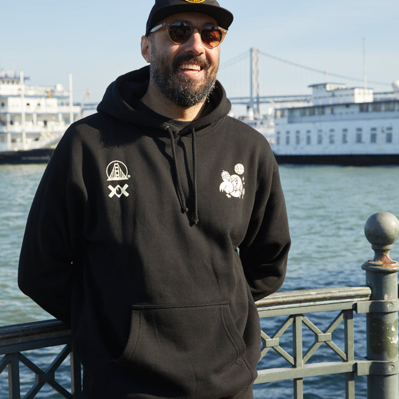 A man wearing a Moon Bridge Pullover Hoodie smiles by the waterfront, with boats and a bridge visible in the background.