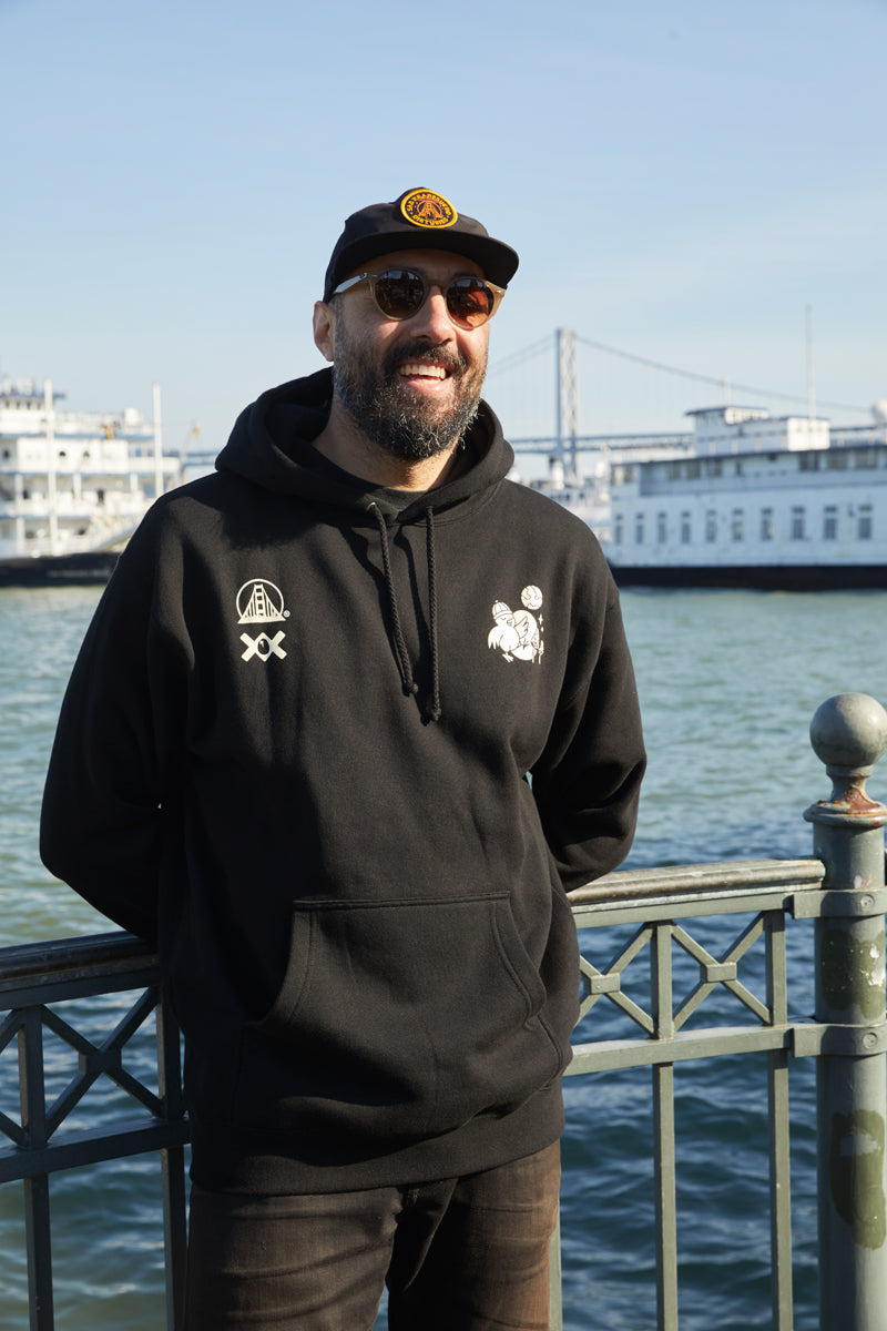 A man wearing a Moon Bridge Pullover Hoodie smiles by the waterfront, with boats and a bridge visible in the background.