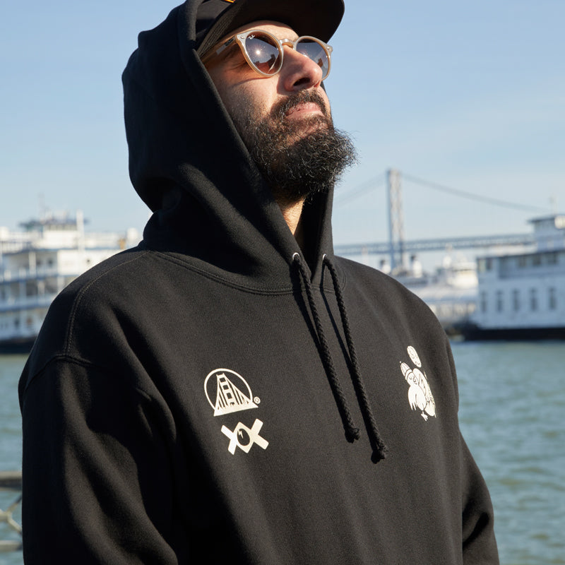 A bearded man in a black Moon Bridge Pullover Hoodie and sunglasses gazes upward by the water, with boats and a bridge behind him.