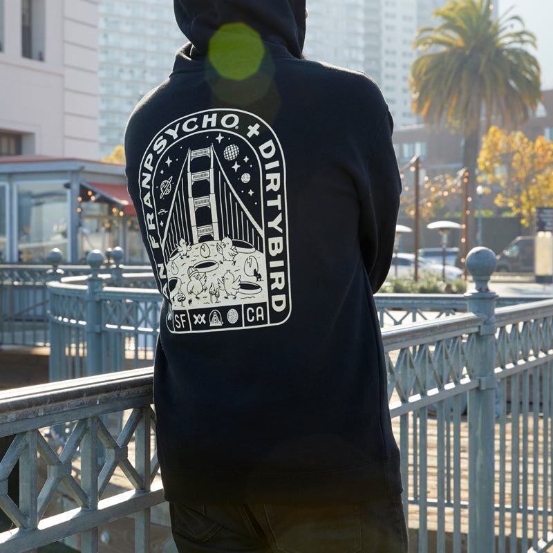 Wearing the Moon Bridge Pullover Hoodie, a person stands by a railing with city buildings and a palm tree in the background.