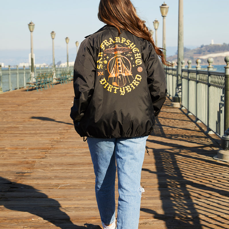 A woman walks on a wooden boardwalk, wearing the Dirty UFO Coaches Jacket from the SFP x Dirtybird collab with blue jeans—a stylish mix of Bay Area streetwear flair.