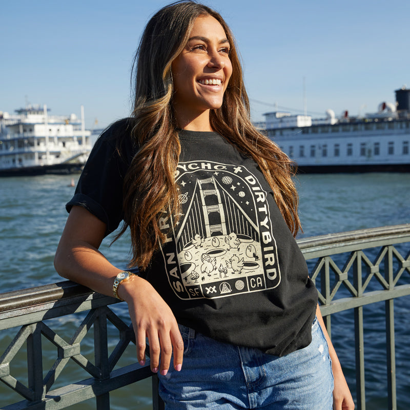 A woman in a Moon Bridge Tee leans on a waterfront railing in San Francisco, with boats in the background.