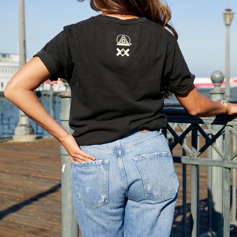 A woman with long hair stands on a San Francisco pier, back to the camera, wearing the Moon Bridge Tee in black and blue jeans.
