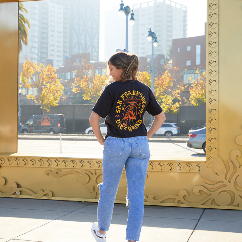 A woman stands outdoors before a large gold frame, wearing jeans and the Dirty UFO Tee—a black graphic t-shirt featuring a San Francisco-inspired hand-printed design that pays homage to Bay Area culture.