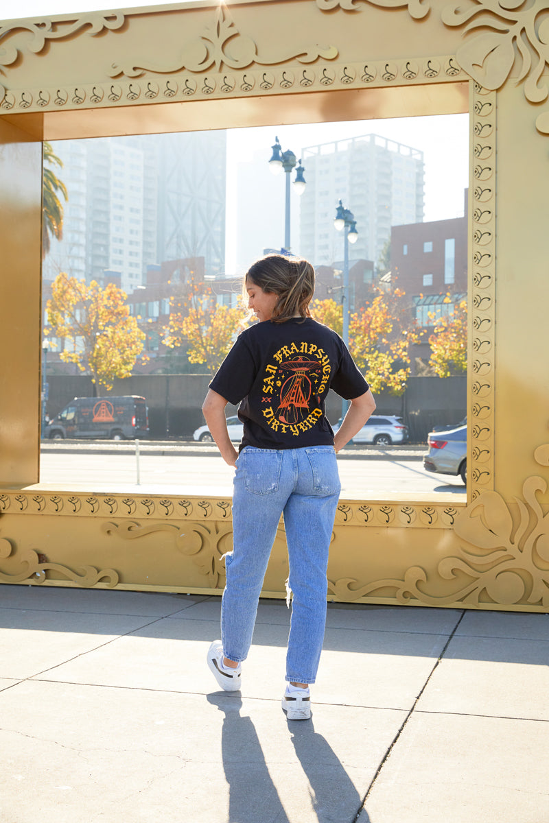 A woman stands outdoors before a large gold frame, wearing jeans and the Dirty UFO Tee—a black graphic t-shirt featuring a San Francisco-inspired hand-printed design that pays homage to Bay Area culture.