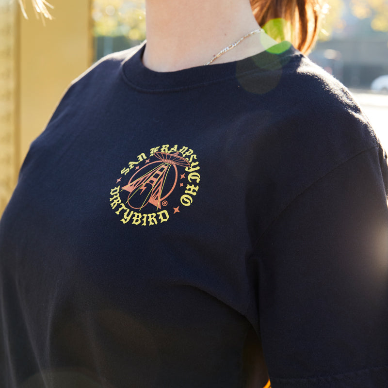 Woman wearing the Dirty UFO Tee—a black shirt with a yellow Dirtybird logo, hand printed in San Francisco—celebrates Bay Area culture with a sun flare in the background.