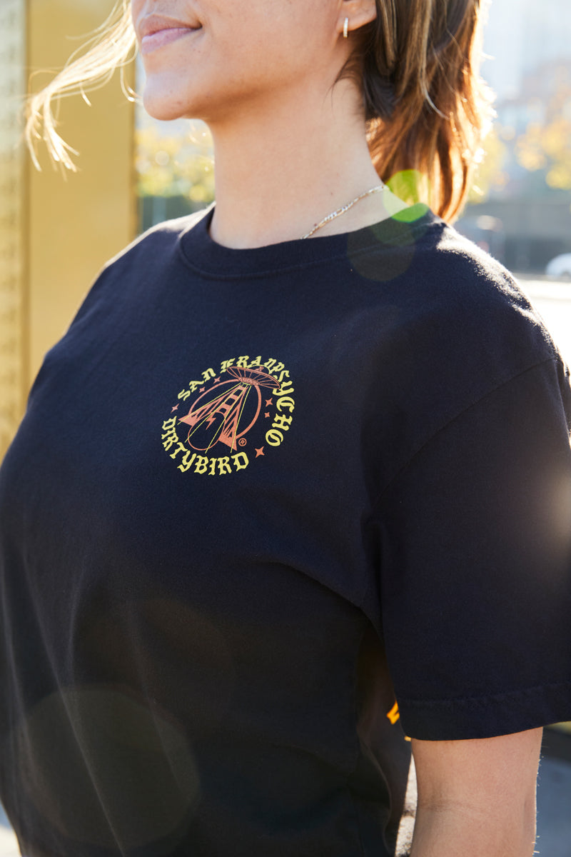 Woman wearing the Dirty UFO Tee—a black shirt with a yellow Dirtybird logo, hand printed in San Francisco—celebrates Bay Area culture with a sun flare in the background.