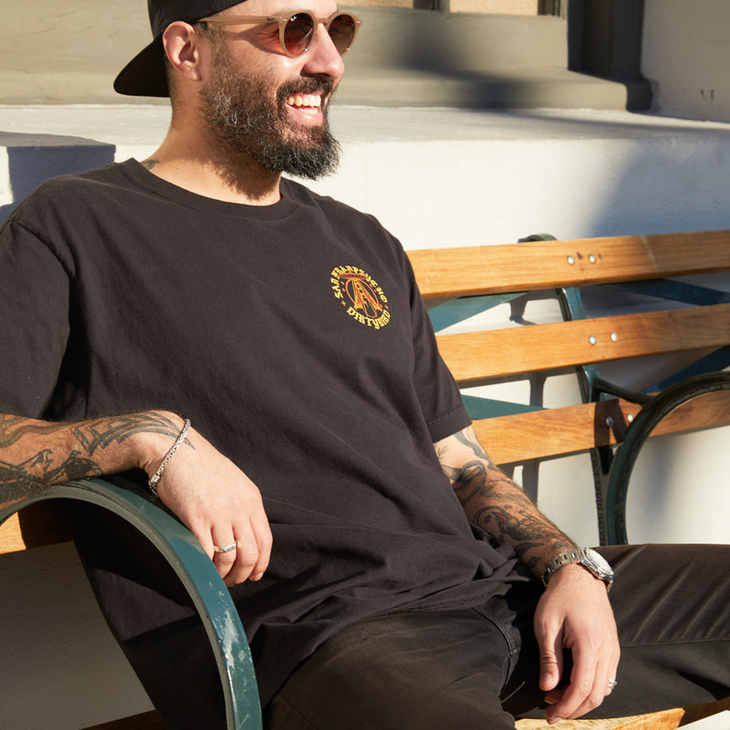 Man with a beard and sunglasses smiles while sitting on a bench in the sunlight, sporting the Dirty UFO Tee and a cap—a perfect nod to Bay Area culture.