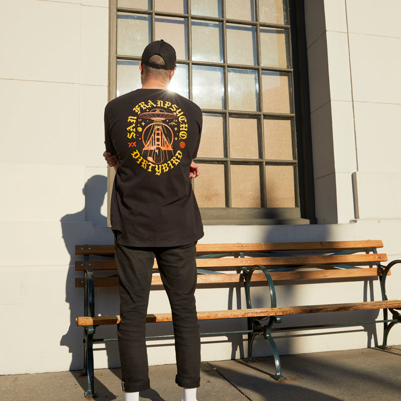A person in a black Dirty UFO Tee and cap stands by a bench outside, facing a sunny window—a scene reflecting Bay Area culture and the creative spirit behind San Francisco hand-printed apparel.
