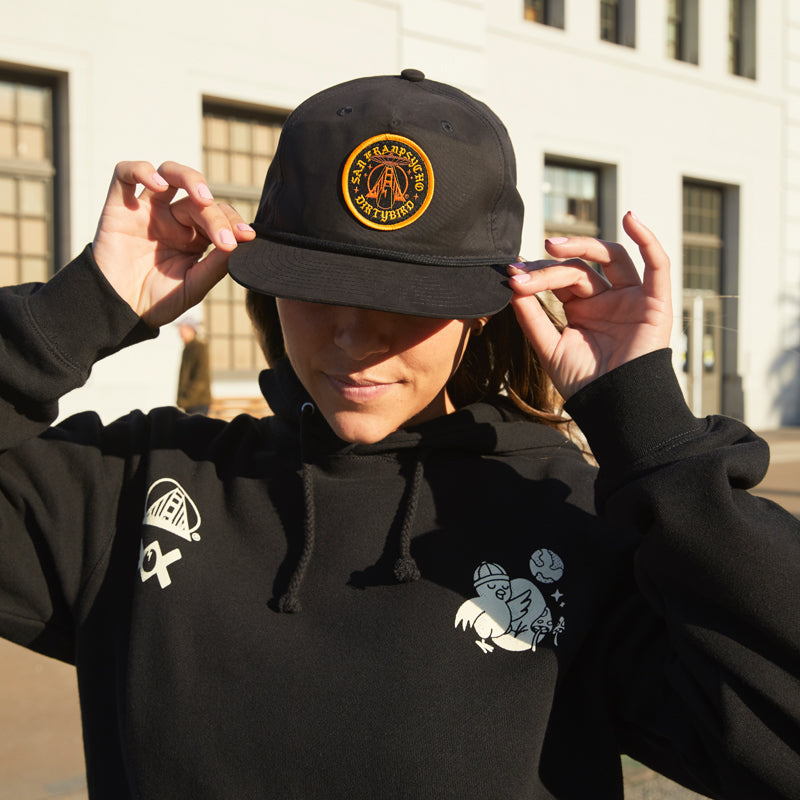A woman in a black hoodie and Dirty UFO Hat stands outside, adjusting her cap with a white building behind her—a nod to Bay Area culture.