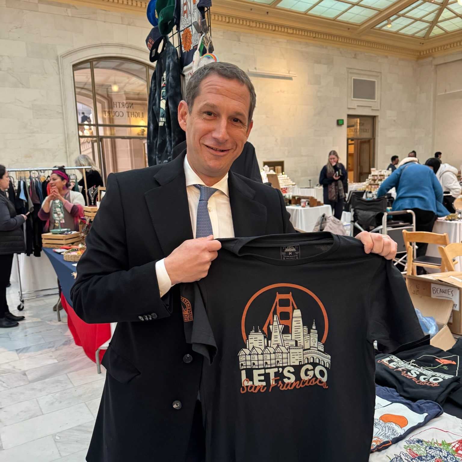 A man in a suit smiles while holding a Let's Go San Francisco Tee at an indoor market or fair.
