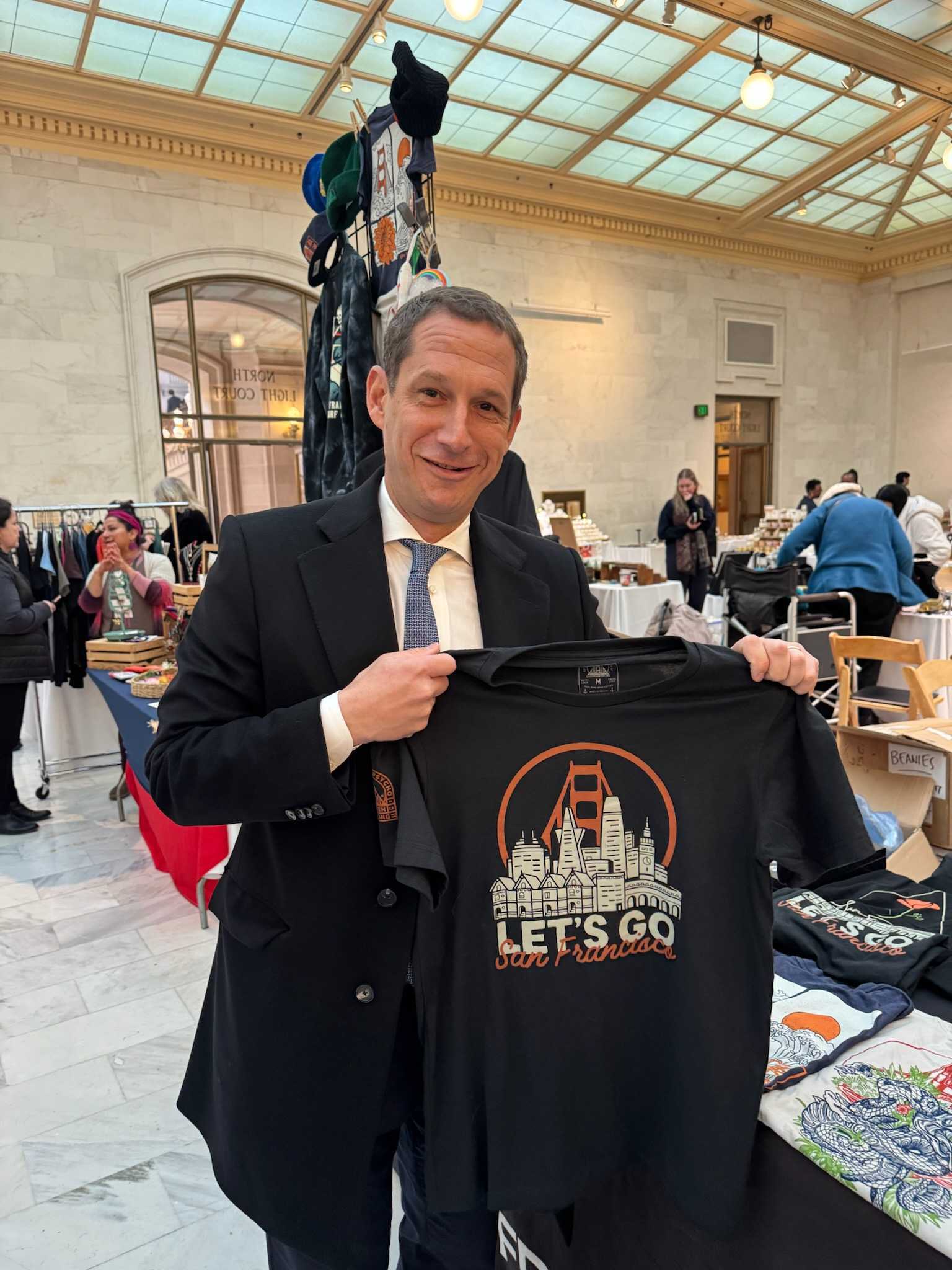 A man in a suit smiles while holding a Let's Go San Francisco Tee at an indoor market or fair.