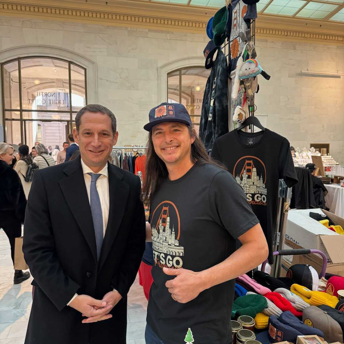 Two men smile in front of a vendor stall selling Let's Go San Francisco Tees and other hand-printed crafts inside a large indoor San Francisco market.