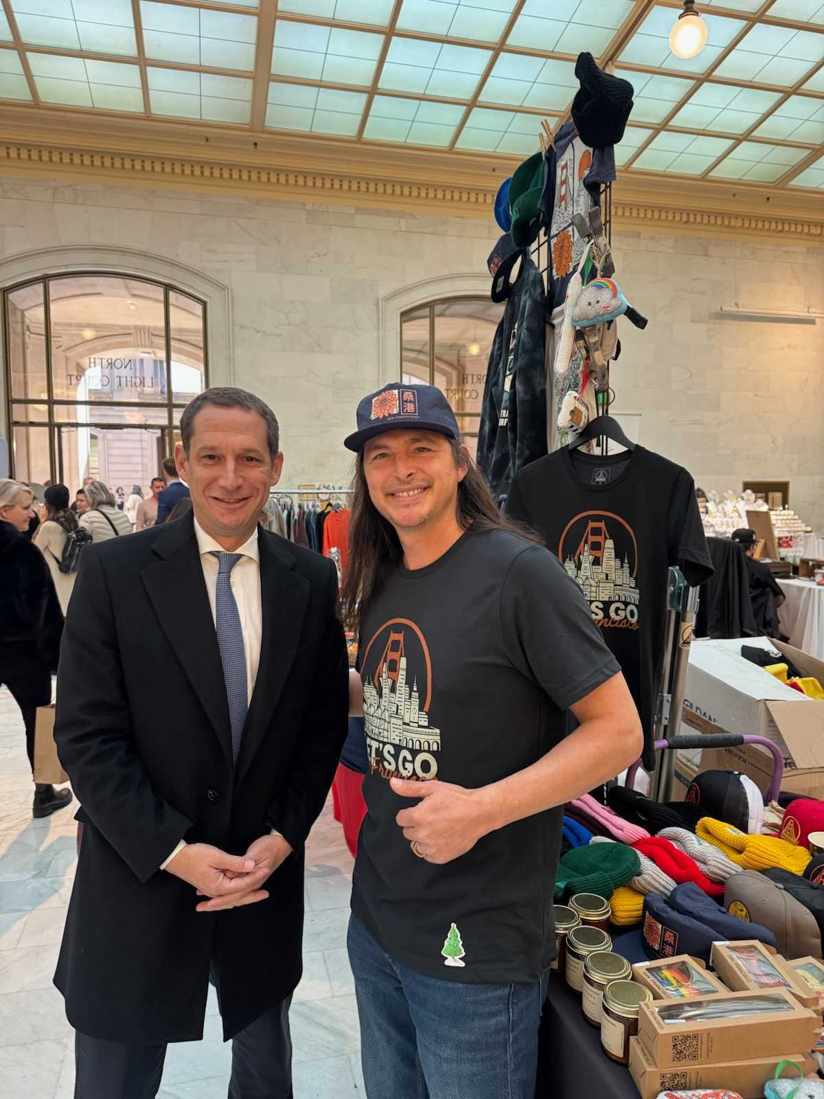 Two men smile in front of a vendor stall selling Let's Go San Francisco Tees and other hand-printed crafts inside a large indoor San Francisco market.