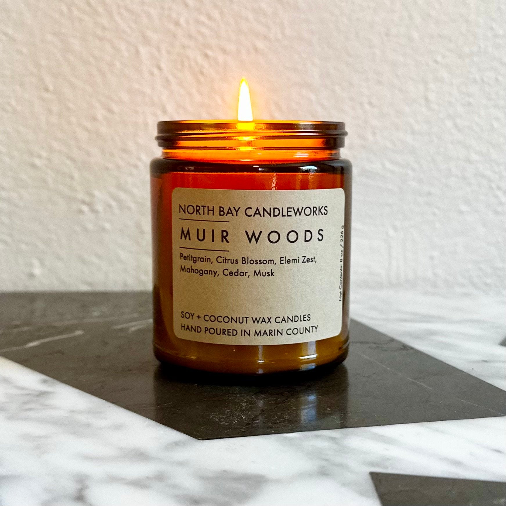 The North Bay Candleworks candle in a brown glass jar, labeled Muir Woods, rests on a marble surface, capturing the tranquil essence of Marin's Bay Area Landmark Line.