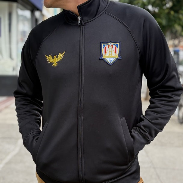 Person wearing the WC x SFP Zip-Up Jacket, a black jacket with gold and colorful embroidered logos, standing outdoors with hands in pockets.