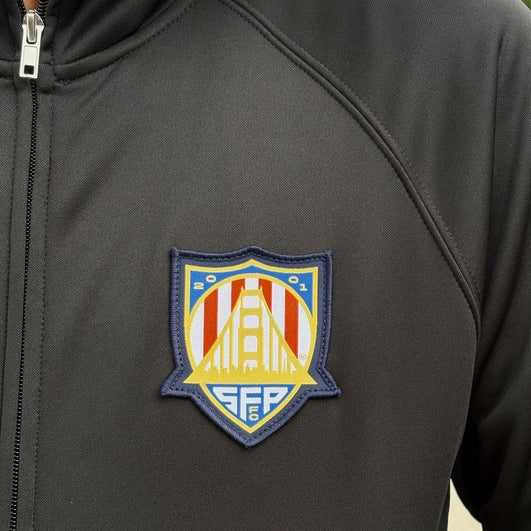 Person wearing the WC x SFP Zip-Up Jacket in black, featuring a colorful shield patch with a bridge and geometric designs.