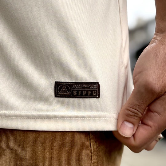 A hand holds the hem of a cream WC x SFP Jersey - Away with an SFPFC patch, next to brown corduroy pants.