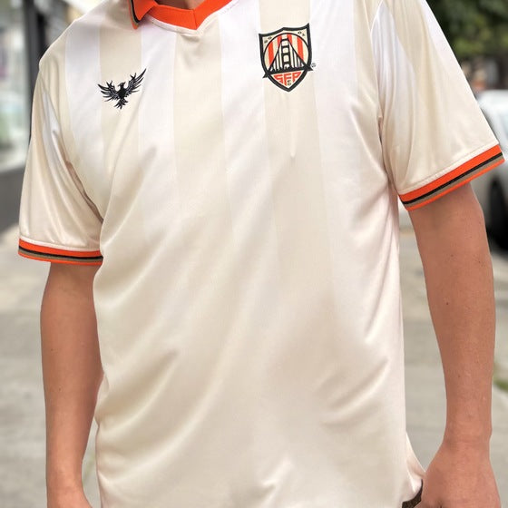 A man is wearing the WC x SFP Jersey - Away, a cream fan-fit top with orange trim and two chest logos, inspired by classic World Cup jerseys, while standing outdoors.