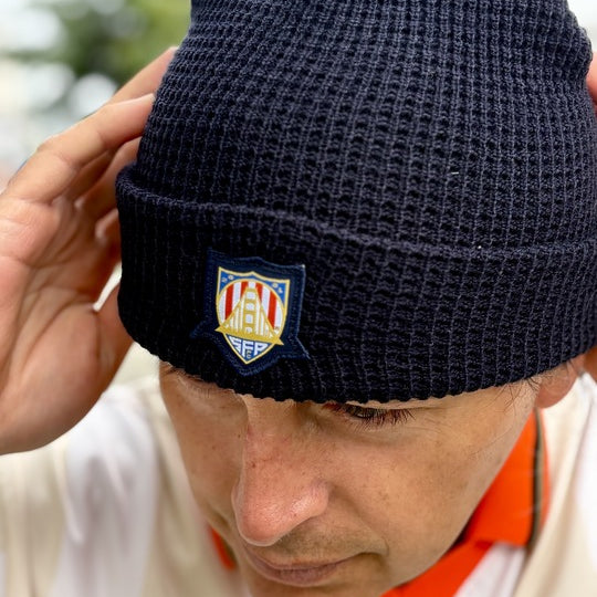 Man in a white shirt and black beanie featuring a crest, looking down and adjusting his WC x SFP Beanie with both hands.