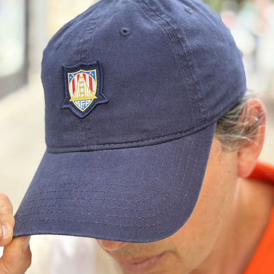 Wearing the WC x SFP Patched Hat with an embroidered crest, a person bows their head and touches the brim—subtly showing support for the US Men's National Team.