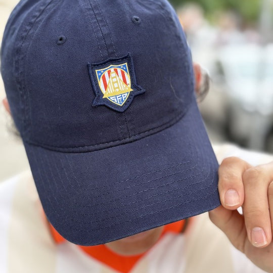 Individual wearing a WC x SFP Patched Hat in navy blue featuring the US Men's National Team patch, paired with a white-striped shirt showcasing the matching shield emblem, while holding the hat’s brim.