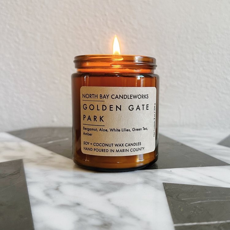 A lit candle in a brown jar rests on a marble surface, its label reading "North Bay Candleworks" with scent notes and candle details. Evoking the essence of the Bay Area, this candle captures the charm found across Marin's Landmark Line.
