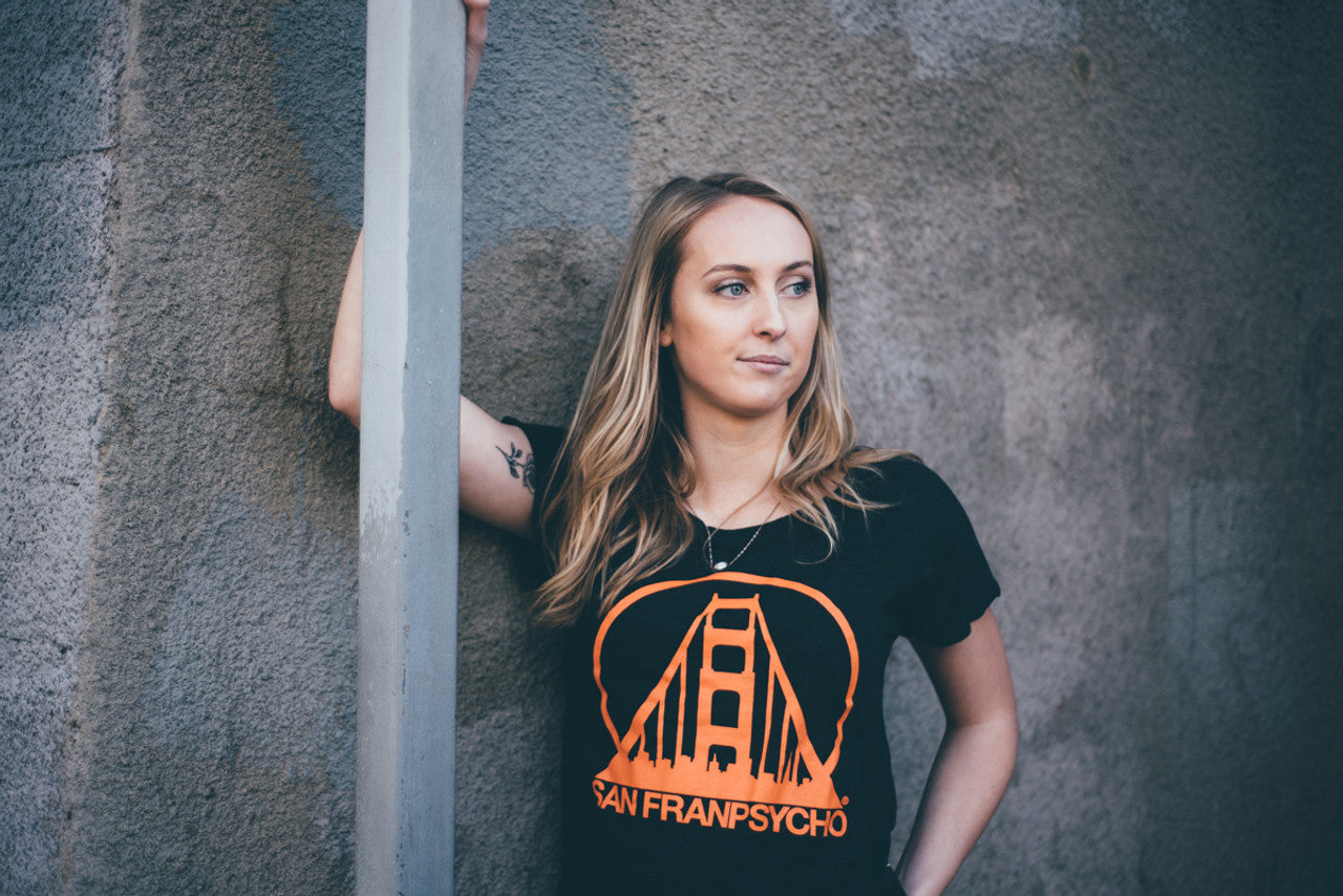 A person leans against a wall, wearing a Charcoal & Orange Dolman t-shirt from San Franpsycho, featuring an orange graphic that is hand-printed in San Francisco.