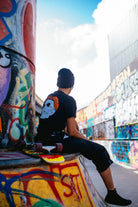 A person sits on a skateboard in a graffiti-covered skate park, wearing a beanie and a Black & Orange Street Skull Tee made from soft Airlume cotton.