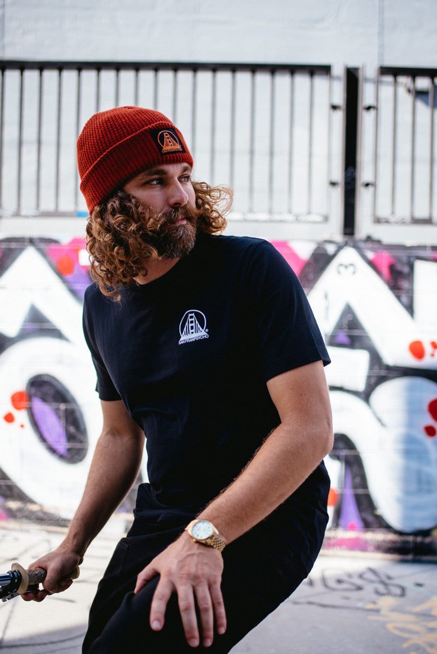 A man with curly hair, donning a red beanie and a Black & Orange Street Skull Tee, stands by a graffiti-covered wall.