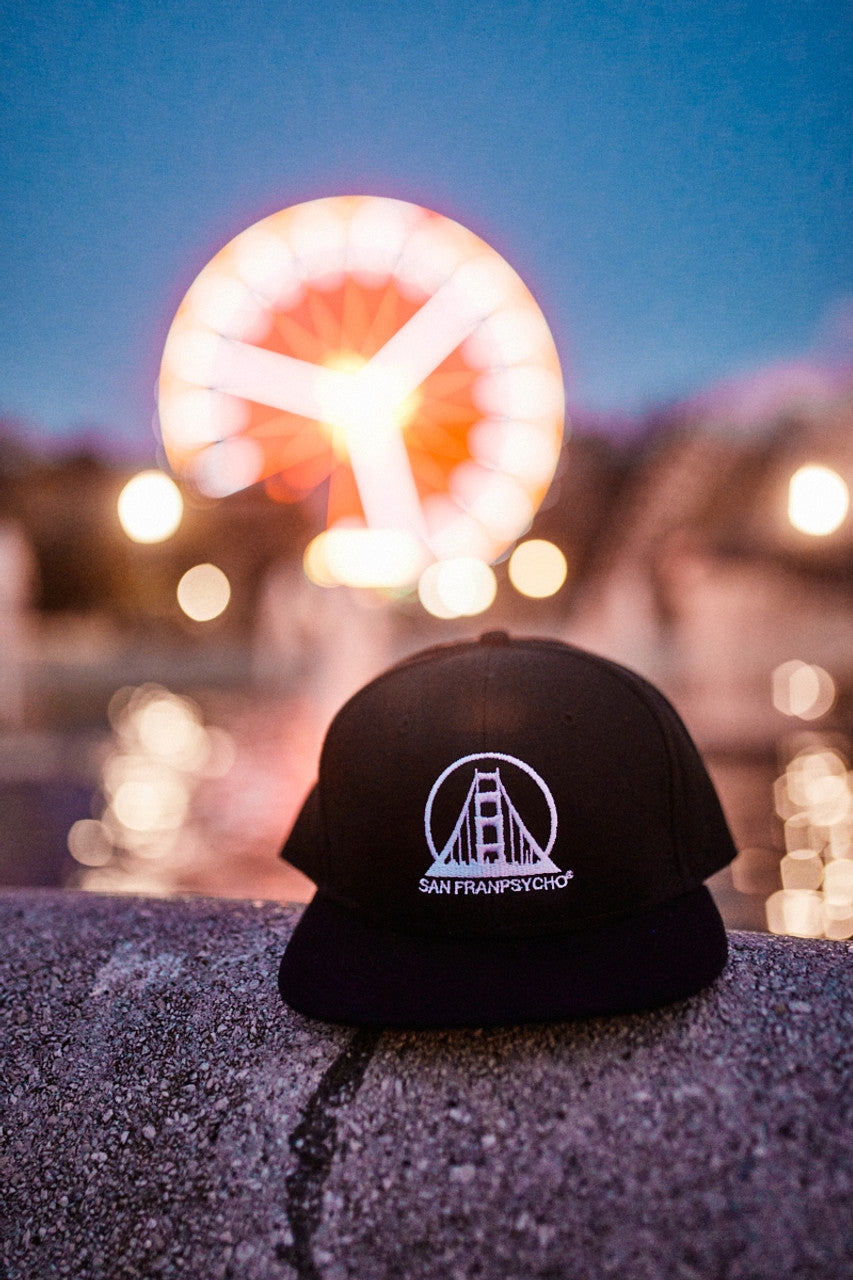 Embroidered Logo Snapback - Black & White cap with a flat brim and SAN FRANPSYCHO logo, set against a blurred Ferris wheel at dusk, featuring snapback adjustment for one size fits most.