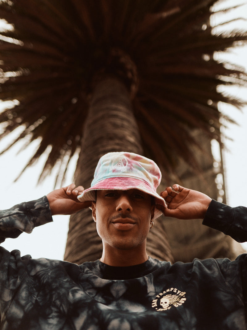A person dressed in a super soft Surf Club Crewneck and a coordinating bucket hat stands under a palm tree, gazing upward, as if anticipating the Skeleton Surf Club hitting the waves.
