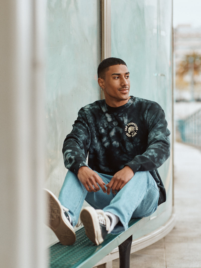 A man wearing a Surf Club Crewneck in a super soft tie-dye pattern sits on a bench, gazing off to the side in a casual outdoor setting.