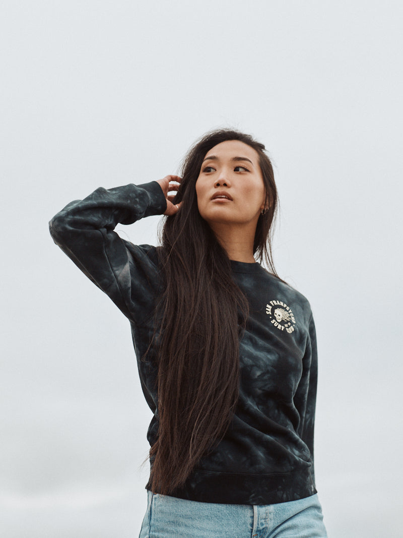 A woman with long hair, dressed in a super soft, dark Surf Club Crewneck sweatshirt and jeans, stands outdoors against a cloudy sky.