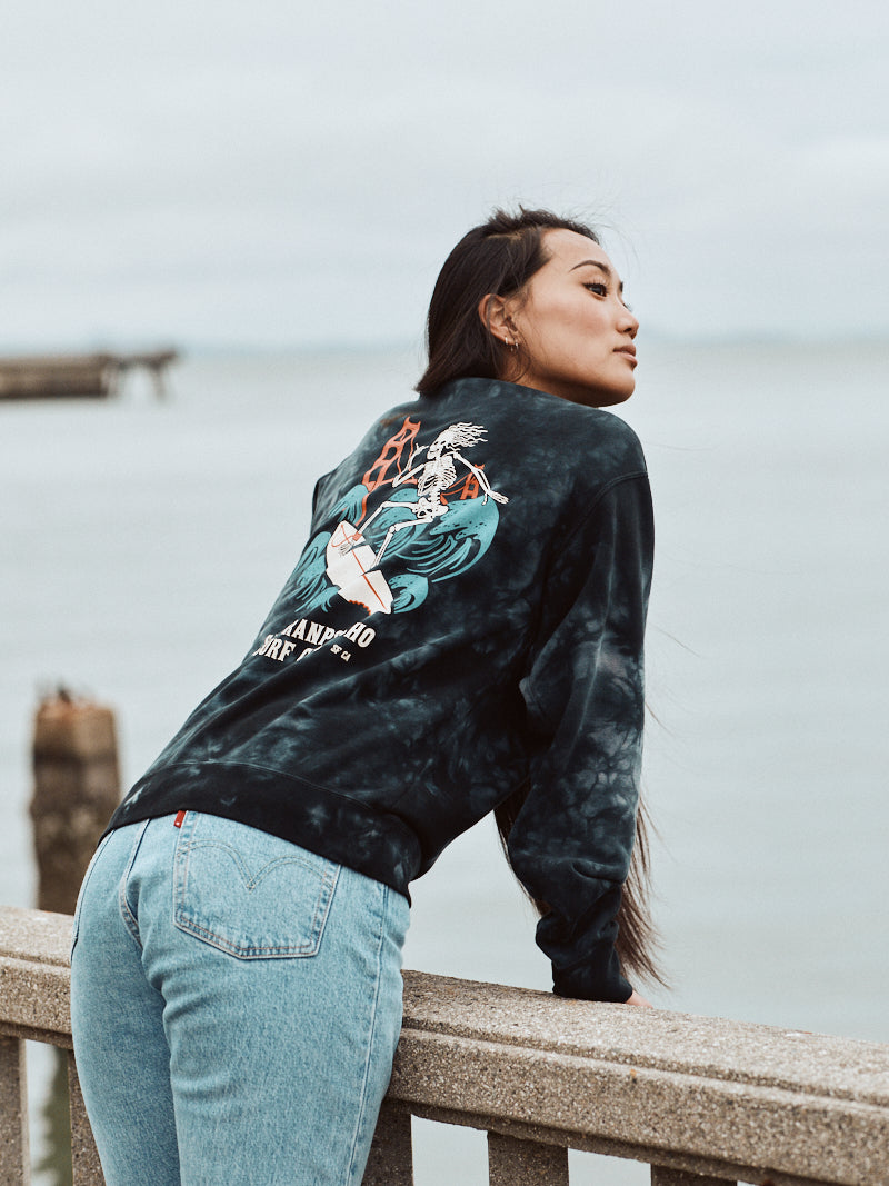 A woman in a black Surf Club Crewneck from the Skeleton line leans on a railing by the sea, her back facing the camera, enjoying the super soft comfort as she gazes at the waves.