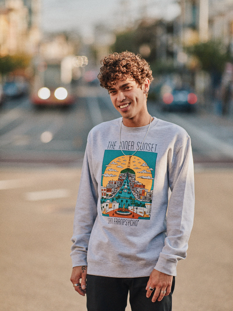A person in an Inner Sunset Crewneck stands with a smile on a bustling street, as buildings and a bus blur in the background.