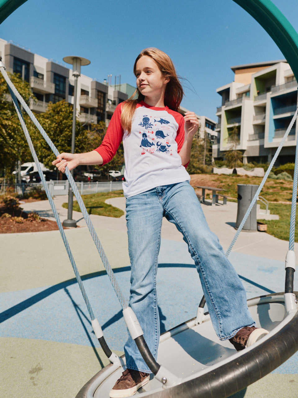 Wearing a Youth Ocean Club Baseball Tee and jeans, a person stands confidently on a playground swing, dreaming of adventurous times with the Kids Ocean Club.