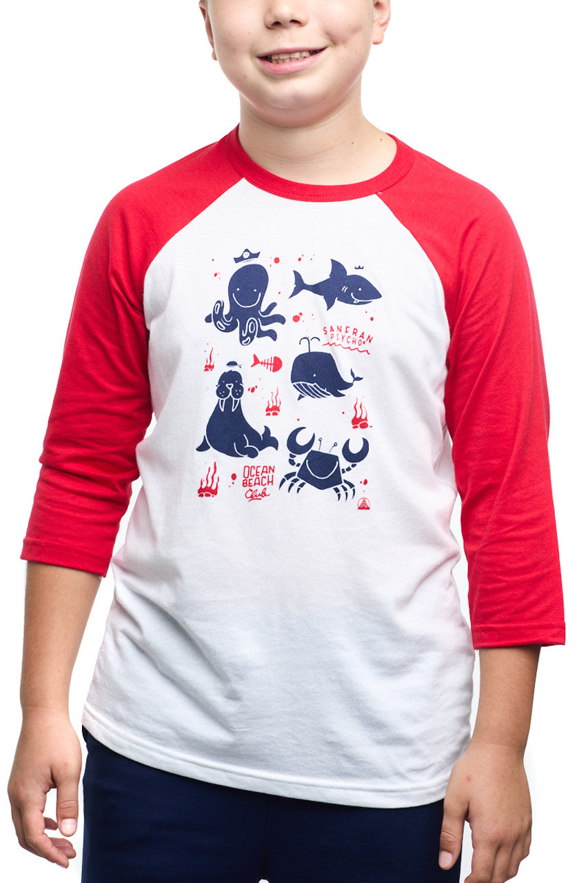 A child wears the Youth Ocean Club Baseball Tee, a fun red and white shirt with cartoon sea creatures and Since Ocean Beach text. Made from 100% cotton, its perfect for young ocean club fans seeking comfort.