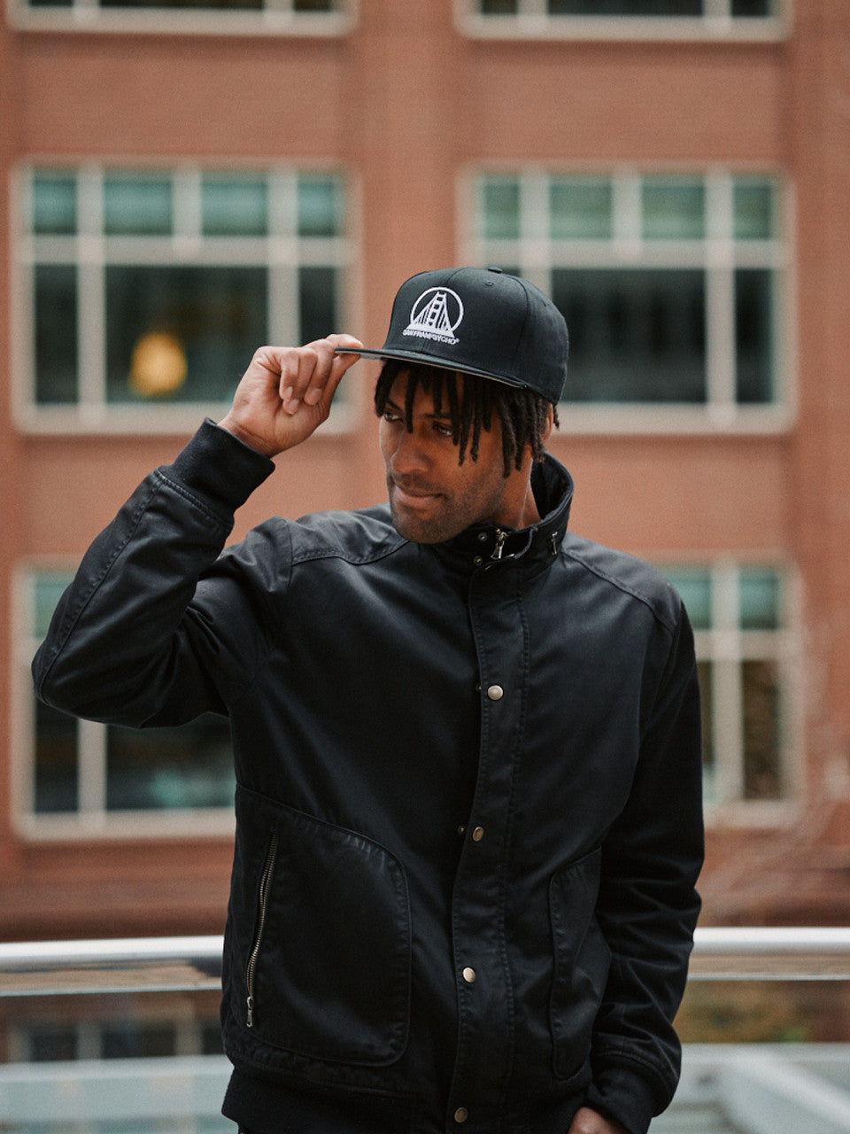 A man wearing an Embroidered Logo Snapback - Black & White stands in front of a brick building, adjusting the brim of his cap.