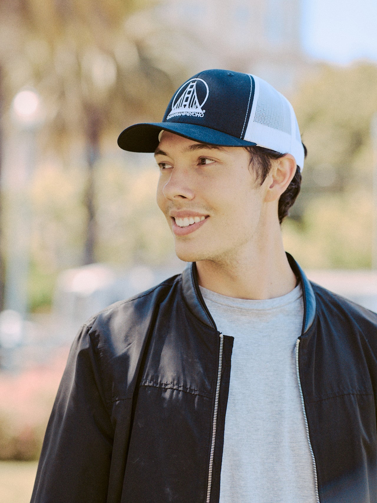 A smiling person wearing the Embroidered Logo Trucker Hat in black and white, accompanied by a jacket, stands outdoors against a serene backdrop of blurred trees.