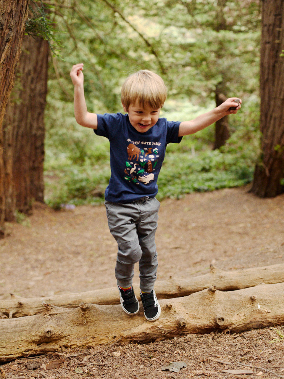 A child joyfully balances on a log in Golden Gate Park, wearing a Youth Golden Gate Park Animals Navy Tee and gray pants, their laughter echoing through the forest.