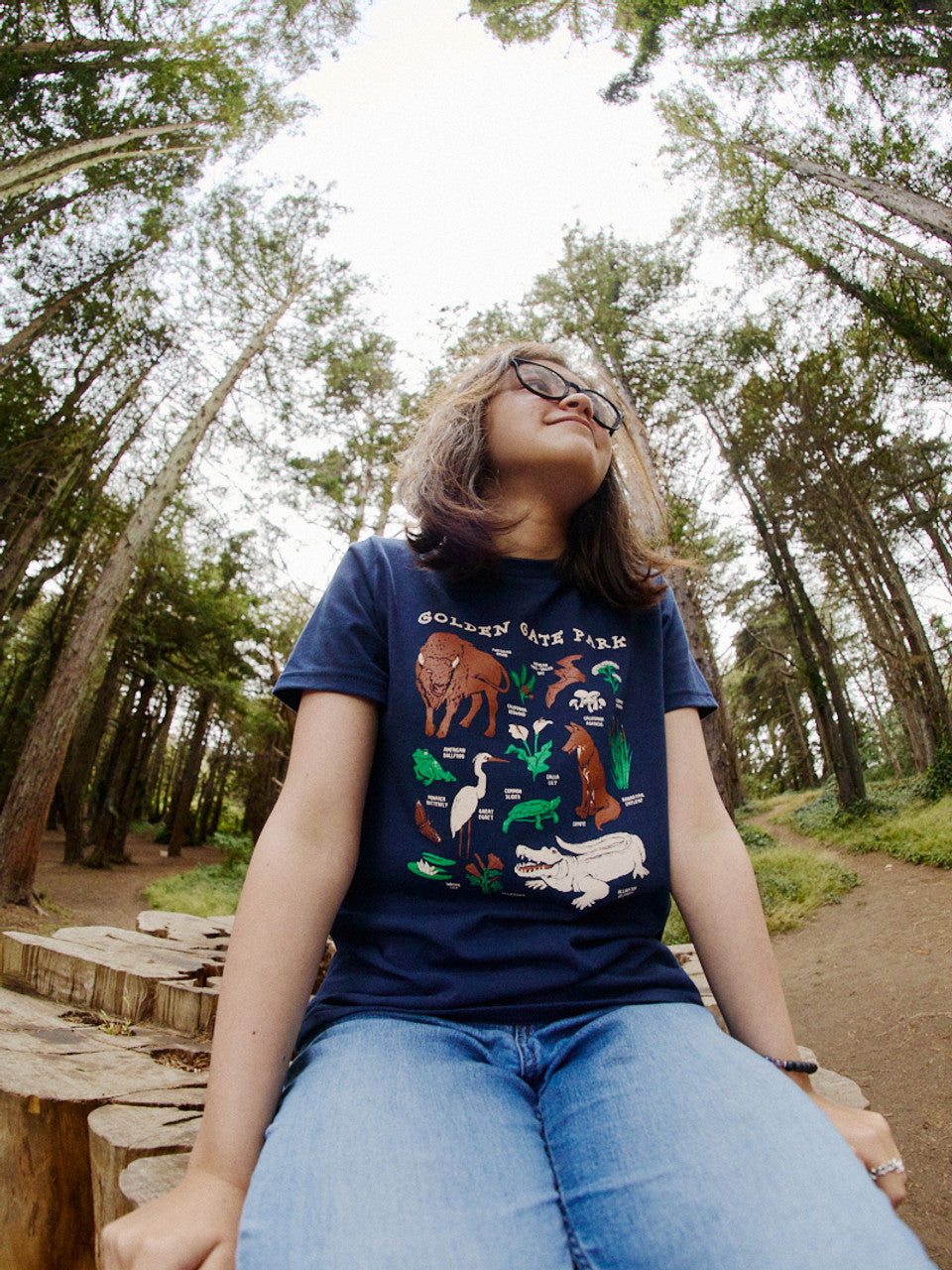 Wearing a Youth Golden Gate Park Animals Navy Tee and jeans, a person sits on a wooden log amidst tall trees in Golden Gate Park, gazing upwards.