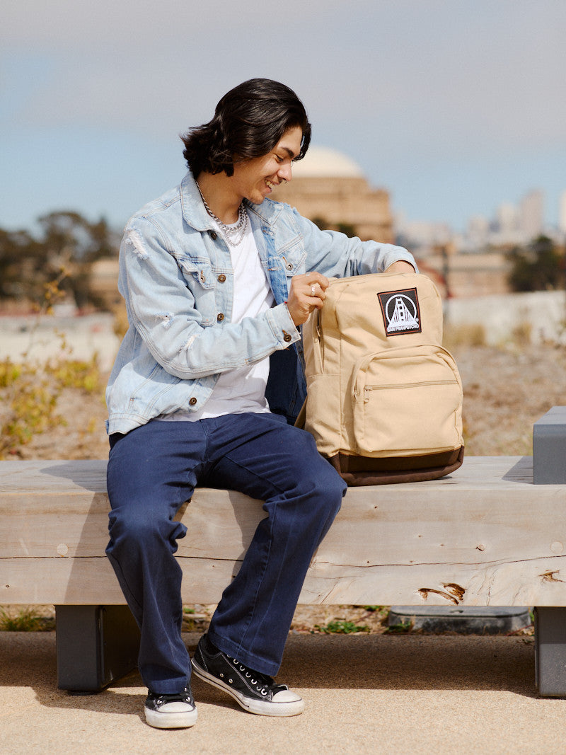 A person sits on a bench, smiling while holding an open Khaki Backpack w/ SFP Patch with leather trims, wearing a denim jacket and blue pants.
