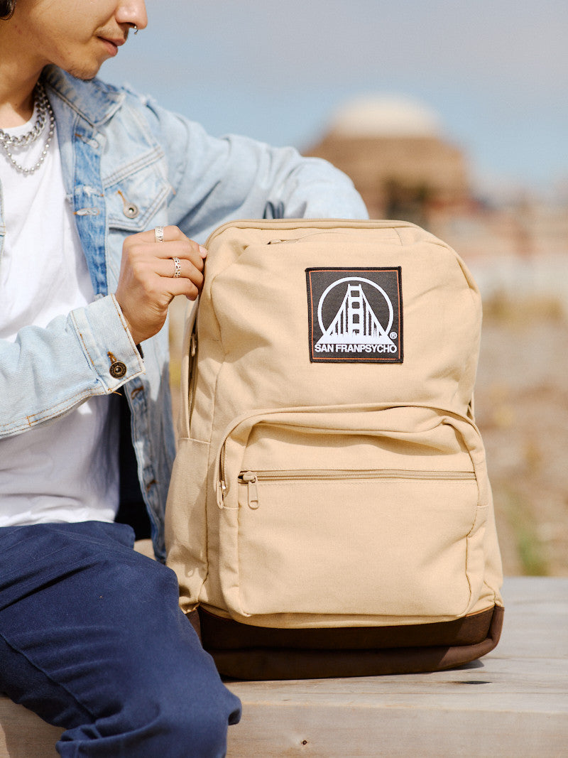 A person clad in jeans and a denim jacket holds the Khaki Backpack w/ SFP Patch, adorned with leather trims, while savoring the outdoor experience.