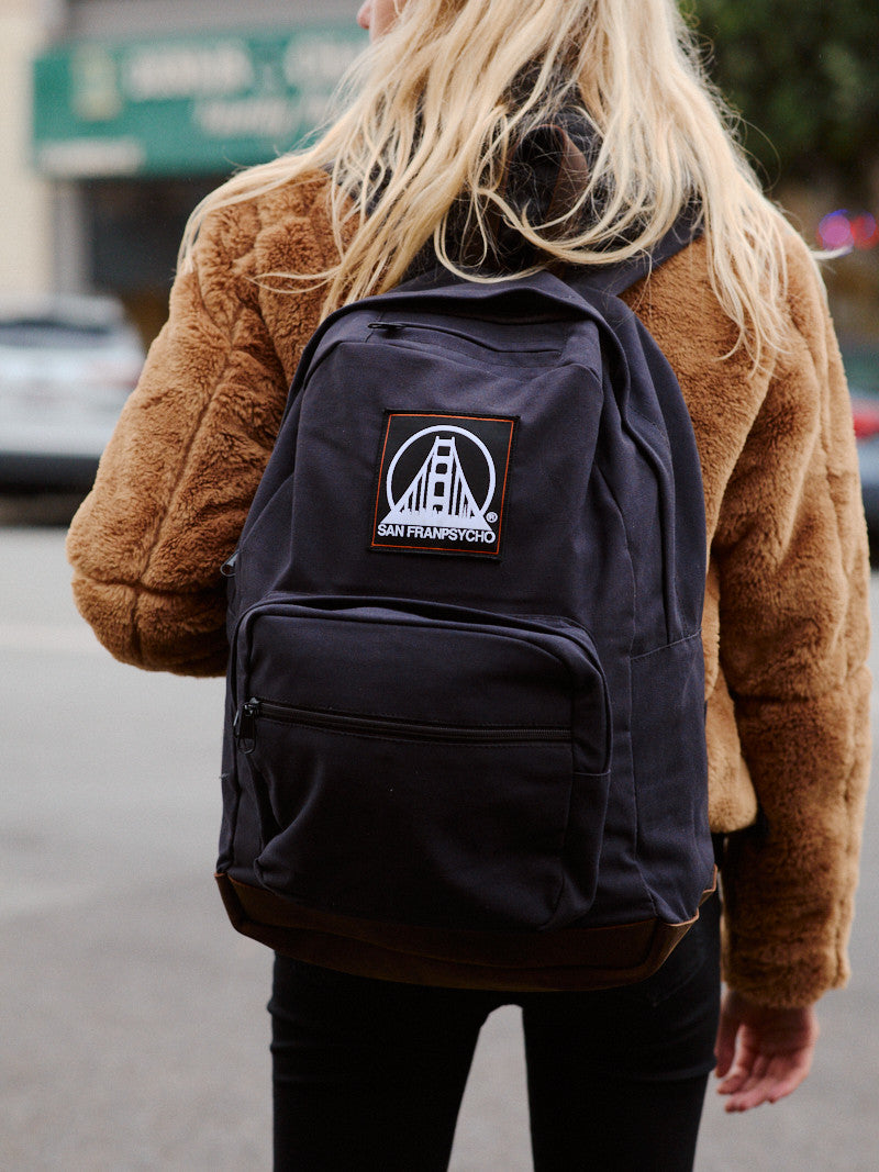 A person with a furry jacket and a Black Backpack w/ SFP Patch featuring leather trims is standing outdoors.