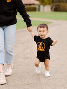 A toddler wearing a Black & Orange Logo Onesie walks while holding an adult's hand along a sunny path.