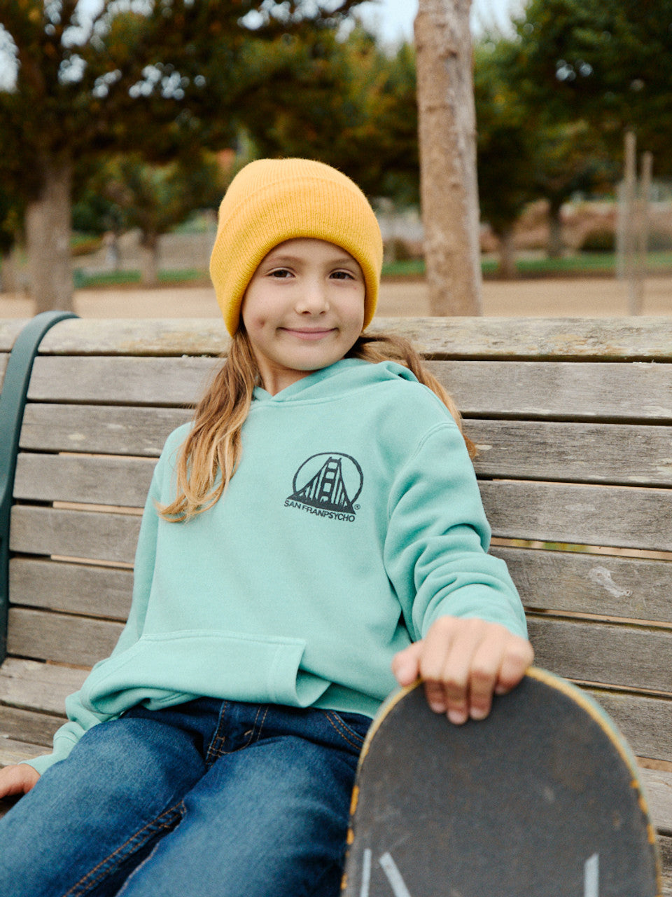 A young person in a yellow beanie and Youth Mint Scribble Logo Pullover Hoodie sits on a bench with a skateboard, effortlessly blending casual style.