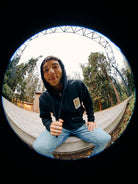 A person in a Black Muir Woods Patched Zip-Up Hoodie sits on a wooden platform surrounded by trees, all viewed through the intriguing distortion of a fisheye lens. Embracing the spirit of 'Free To Wander,' this scene captures a moment of quiet reflection in nature.